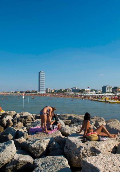 Spiaggia affollata, sole splendente, persone rilassate su scogli.