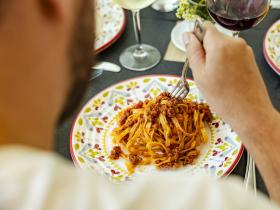 Persona gusta tagliatelle al ragù, atmosfera conviviale con vino e piatti decorati.